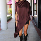 A woman wearing a draped, asymmetrical dark burgundy satin top and black cowboy boots, walking on a covered outdoor walkway.