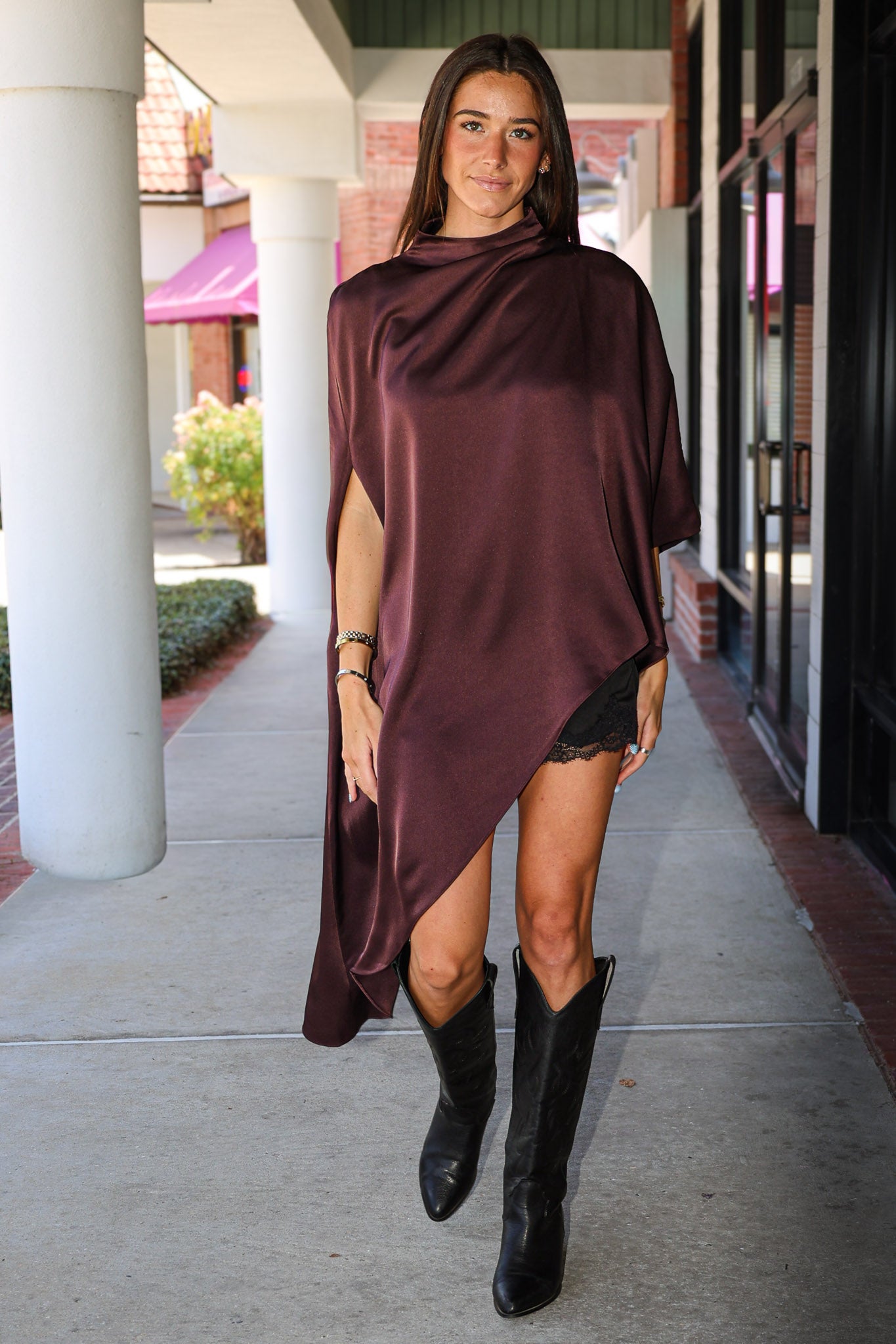 A woman wearing a draped, asymmetrical dark burgundy satin top and black cowboy boots, walking on a covered outdoor walkway.