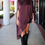 A woman walking outdoors on a sidewalk wearing a draped, high-low satin poncho dress in deep plum purple and black cowboy boots.