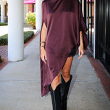 A woman walking outdoors on a sidewalk wearing a draped, high-low satin poncho dress in deep plum purple and black cowboy boots.