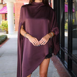 A woman wearing a draped, high-neck, asymmetrical satin poncho in deep plum purple, styled with black cowboy boots, standing on a sidewalk in front of a storefront.