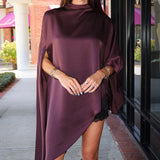 A woman wearing a draped, high-neck, asymmetrical satin poncho in deep plum purple, styled with black cowboy boots, standing on a sidewalk in front of a storefront.
