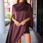 A woman wearing a draped, high-low satin poncho in a deep plum purple color, styled with black shorts and black cowboy boots, standing on a sidewalk.