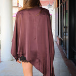 A woman viewed from behind wearing a draped, asymmetrical mauve-burgundy poncho-style top over a black lace-trimmed mini dress, standing on a covered outdoor walkway.