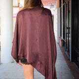 A woman viewed from behind wearing a draped, asymmetrical mauve-burgundy poncho-style top over a black lace-trimmed mini dress, standing on a covered outdoor walkway.