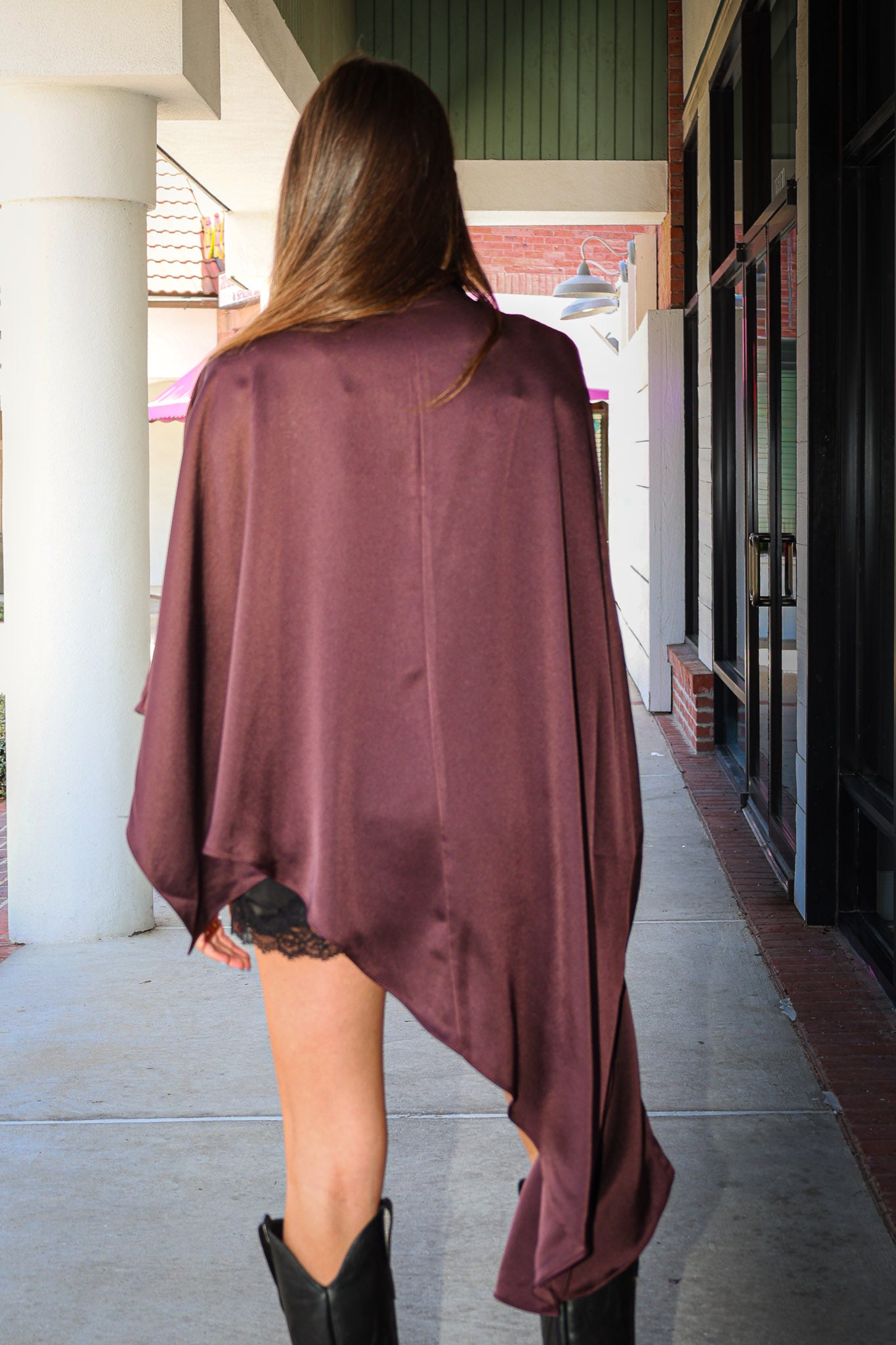 A woman viewed from behind wearing a draped, asymmetrical mauve-burgundy poncho-style top over a black lace-trimmed mini dress, standing on a covered outdoor walkway.