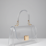 A clear transparent handbag with a curved top handle, gold-tone hardware, and a gold square clasp, photographed on a light gray studio background.