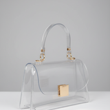 A clear transparent handbag with a curved top handle, gold-tone hardware, and a gold square clasp, photographed on a light gray studio background.