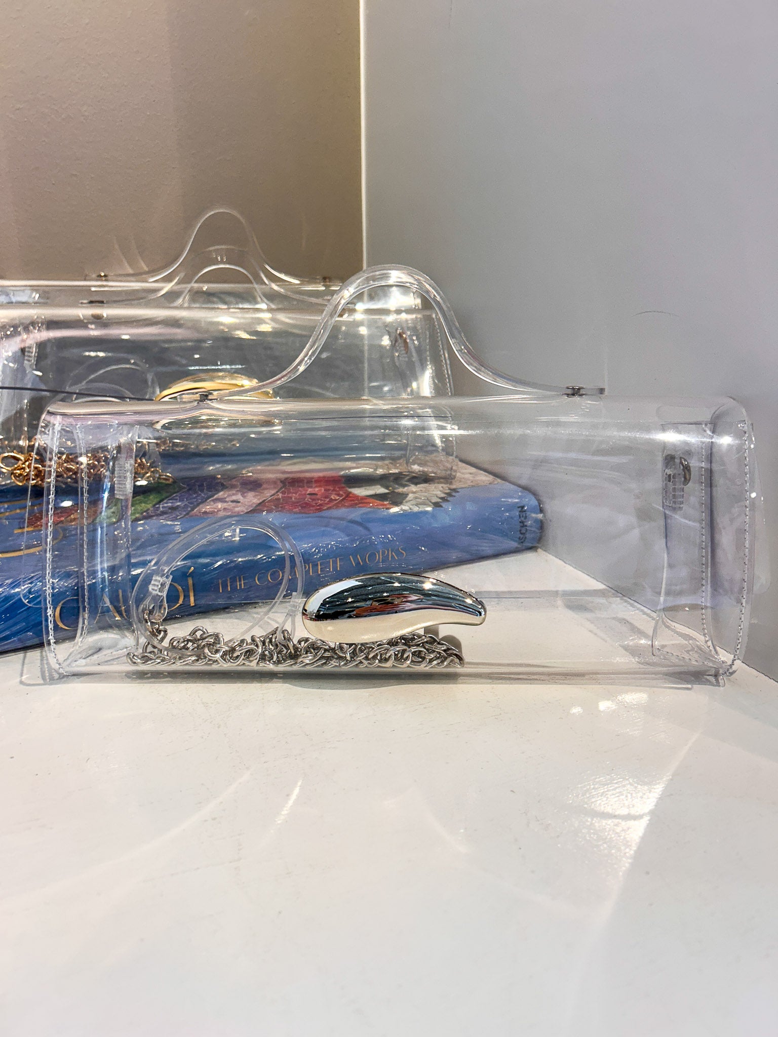 A clear plastic clutch bag with a silver metallic handle, resting on a white surface with a blue book in the background.