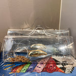 A clear acrylic clutch bag with a transparent handle, shown resting on top of a blue book. Inside the clutch are several gold and silver rings, and the background features a neutral beige wall.