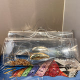 A clear acrylic clutch bag with a transparent handle, shown resting on top of a blue book. Inside the clutch are several gold and silver rings, and the background features a neutral beige wall.