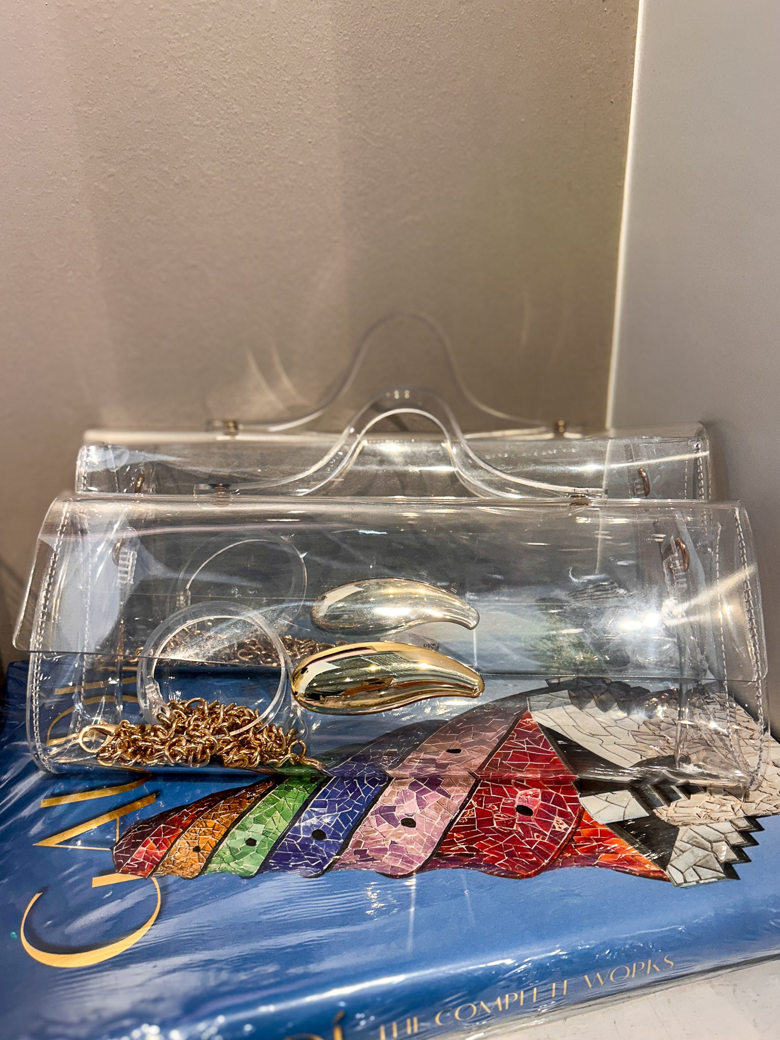 A clear acrylic clutch bag with a transparent handle, shown resting on top of a blue book. Inside the clutch are several gold and silver rings, and the background features a neutral beige wall.