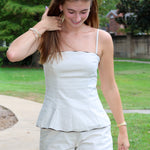 A woman wearing a beige two-piece matching set consisting of a sleeveless camisole top with thin straps and matching shorts, standing outdoors on a paved path with green grass and trees in the background.