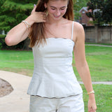 A woman wearing a beige two-piece matching set consisting of a sleeveless camisole top with thin straps and matching shorts, standing outdoors on a paved path with green grass and trees in the background.