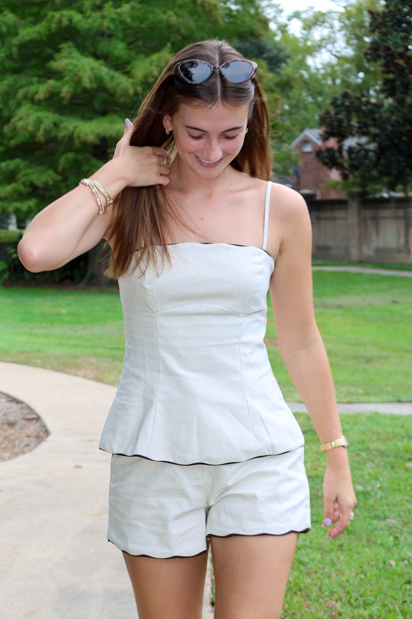 A woman wearing a beige two-piece matching set consisting of a sleeveless camisole top with thin straps and matching shorts, standing outdoors on a paved path with green grass and trees in the background.