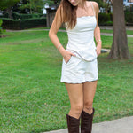 A woman stands outdoors on a concrete path in a grassy park area, wearing a beige matching short set with brown piping with scalloped edges and dark brown knee-high leather boots.