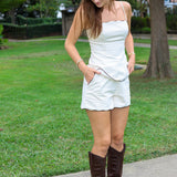 A woman stands outdoors on a concrete path in a grassy park area, wearing a beige matching short set with brown piping with scalloped edges and dark brown knee-high leather boots.