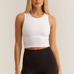 A woman modeling a white ribbed sleeveless crop top paired with black leggings, standing against a plain light beige studio background.