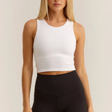A woman modeling a white ribbed sleeveless crop top paired with black leggings, standing against a plain light beige studio background.