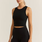 A woman modeling a black ribbed sleeveless tank top and matching black leggings against a plain light beige studio background.