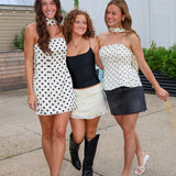 Three women walking together outdoors on a paved area, each wearing a matching white halter top with black polka dots in different styling combinations: one with a white mini skirt, one in a black camisole and white skirt, and one in a black mini skirt.