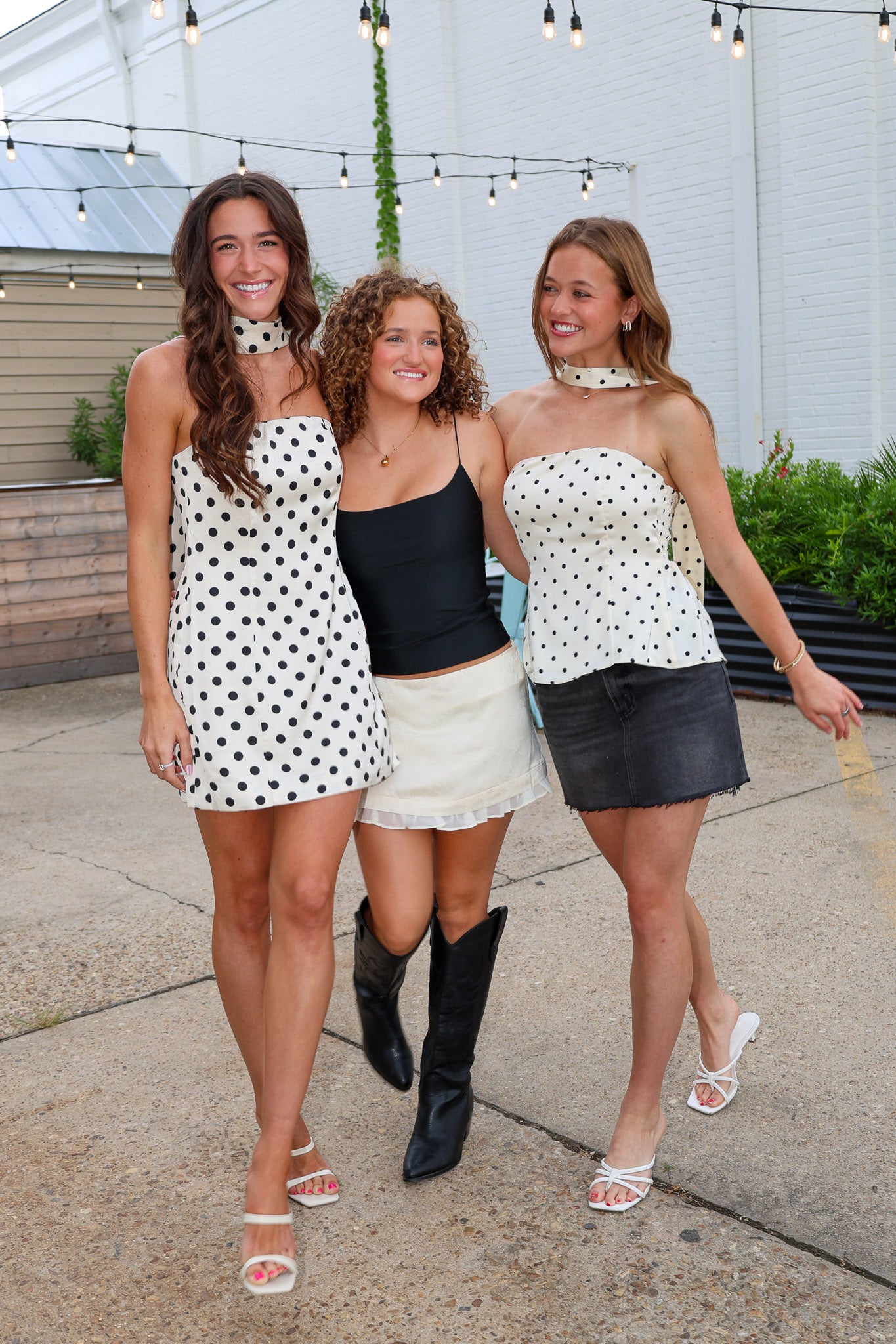 Three women walking together outdoors on a paved area, each wearing a matching white halter top with black polka dots in different styling combinations: one with a white mini skirt, one in a black camisole and white skirt, and one in a black mini skirt.