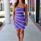 A woman modeling a strapless purple dress with white wavy trim, walking on a covered outdoor walkway.