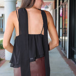 A woman shown from behind wearing a black sleeveless top with a draped open back and layered ruffle detail, paired with a brown skirt, in an outdoor walkway setting.