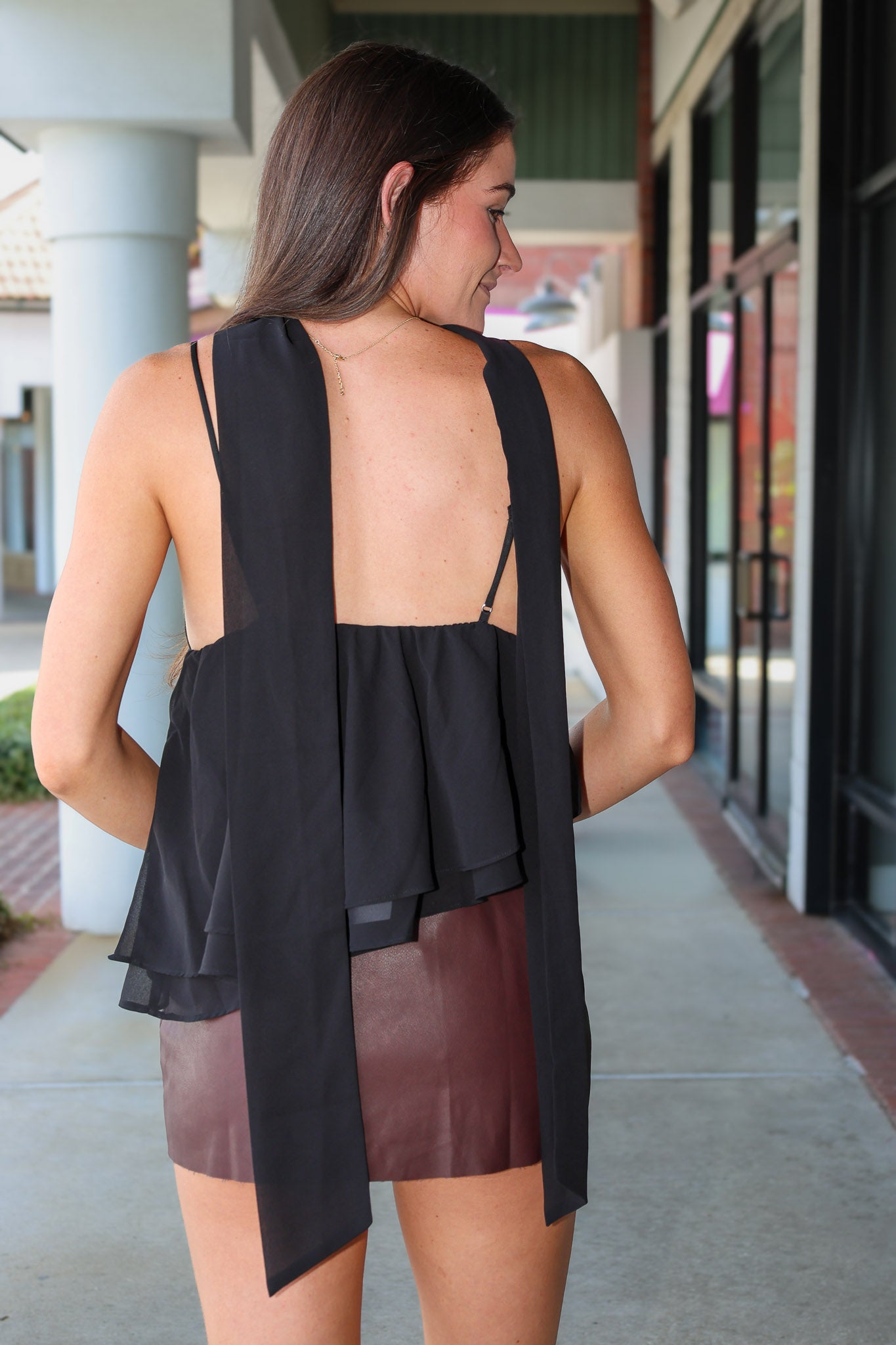 A woman shown from behind wearing a black sleeveless top with a draped open back and layered ruffle detail, paired with a brown skirt, in an outdoor walkway setting.