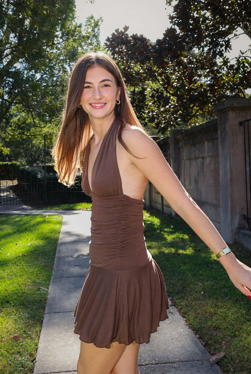 A woman wearing a brown halter-neck mini dress with ruching and a flared skirt, standing outdoors on a paved path with trees and a fence in the background.