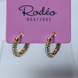 A pair of small gold hoop earrings covered with small clear rhinestones, hanging from a pink display card printed with the brand name 'Rodéo Boutique'.