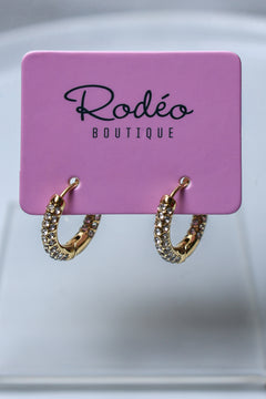 A pair of small gold hoop earrings covered with small clear rhinestones, hanging from a pink display card printed with the brand name 'Rodéo Boutique'.