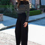Woman wearing a striped sweater and black pants standing on a brick path with a building in the background.