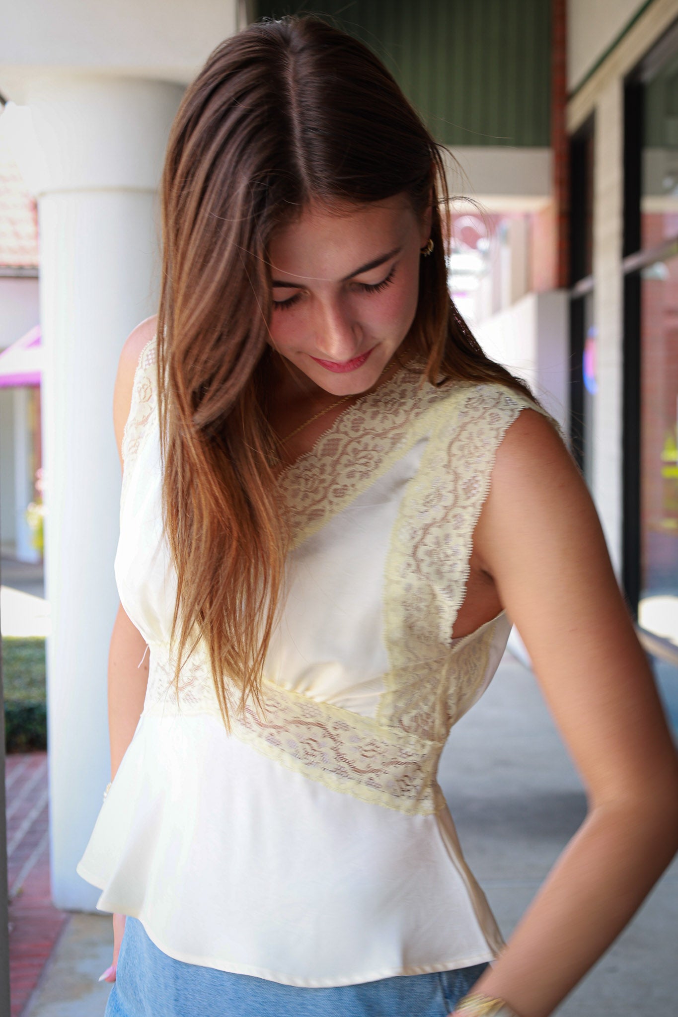 sleeveless cream-colored blouse with wide lace trim and a peplum hem, paired with blue jeans, standing in an outdoor walkway.