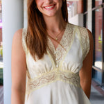  sleeveless cream-colored blouse with wide lace trim and a peplum hem, paired with blue jeans, standing in an outdoor walkway.
