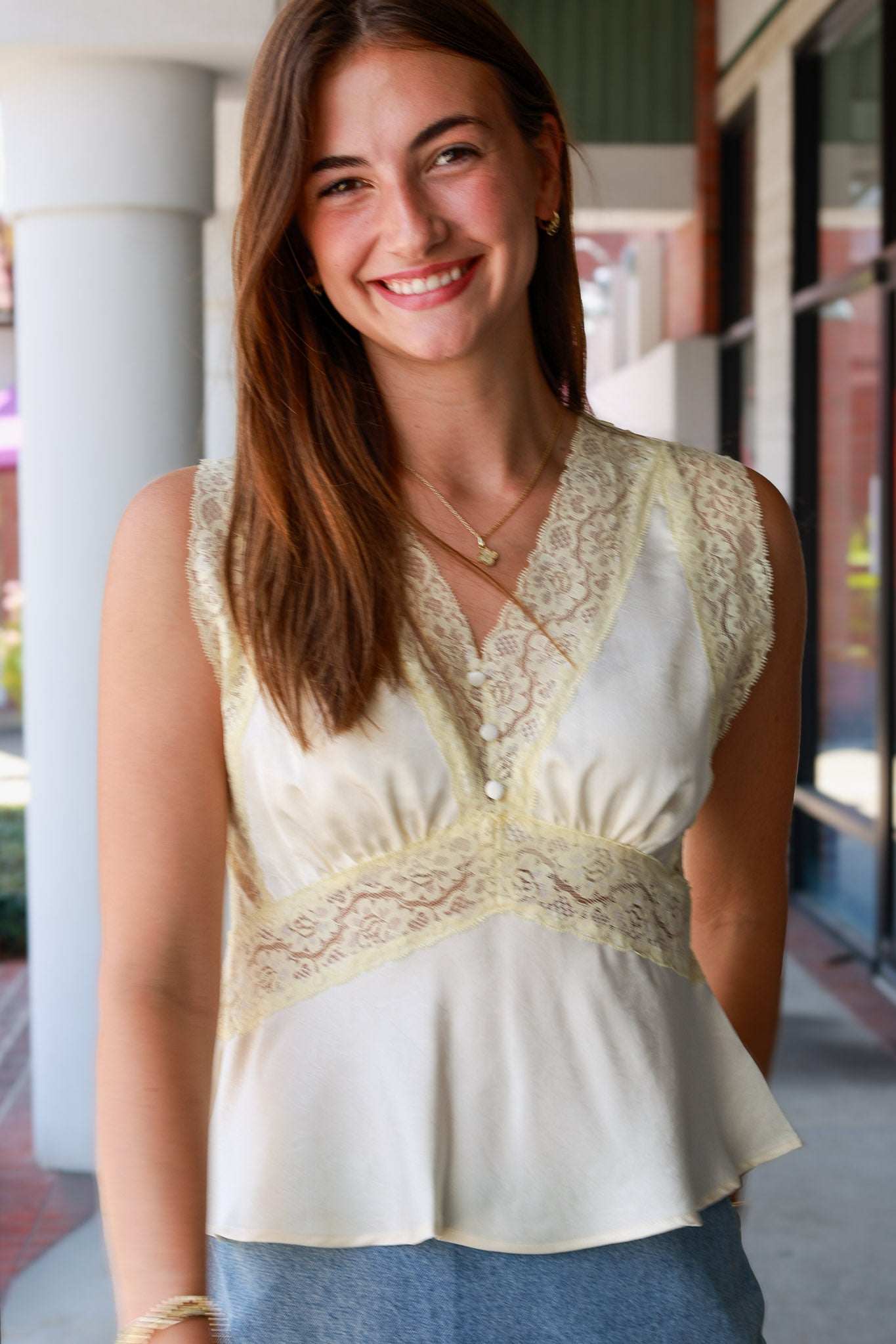  sleeveless cream-colored blouse with wide lace trim and a peplum hem, paired with blue jeans, standing in an outdoor walkway.