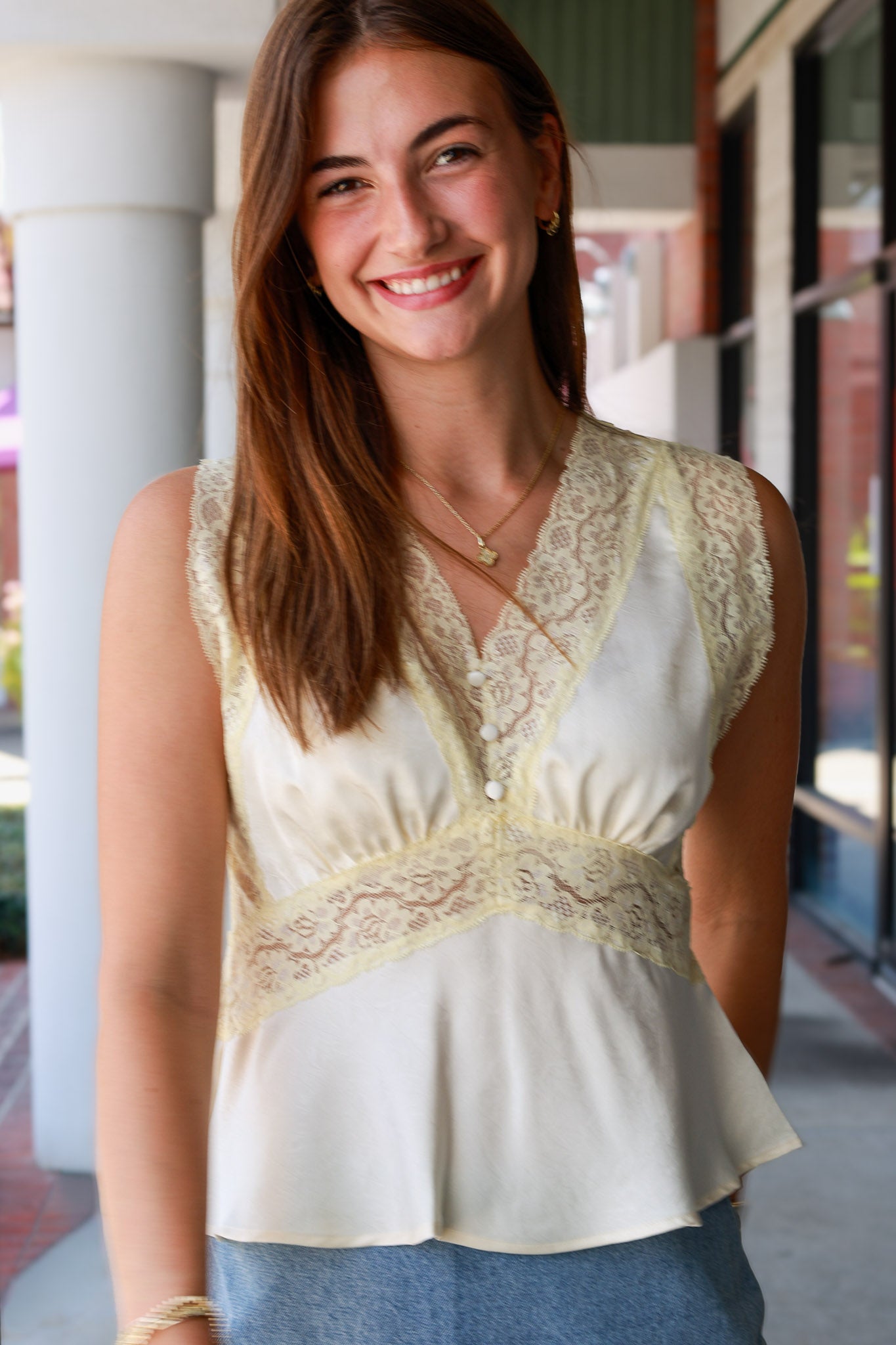  sleeveless cream-colored blouse with wide lace trim and a peplum hem, paired with blue jeans, standing in an outdoor walkway.