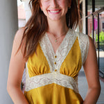 A smiling young woman wearing a sleeveless mustard yellow top with cream lace trim and a buttoned V-neckline, photographed outdoors on a covered walkway.