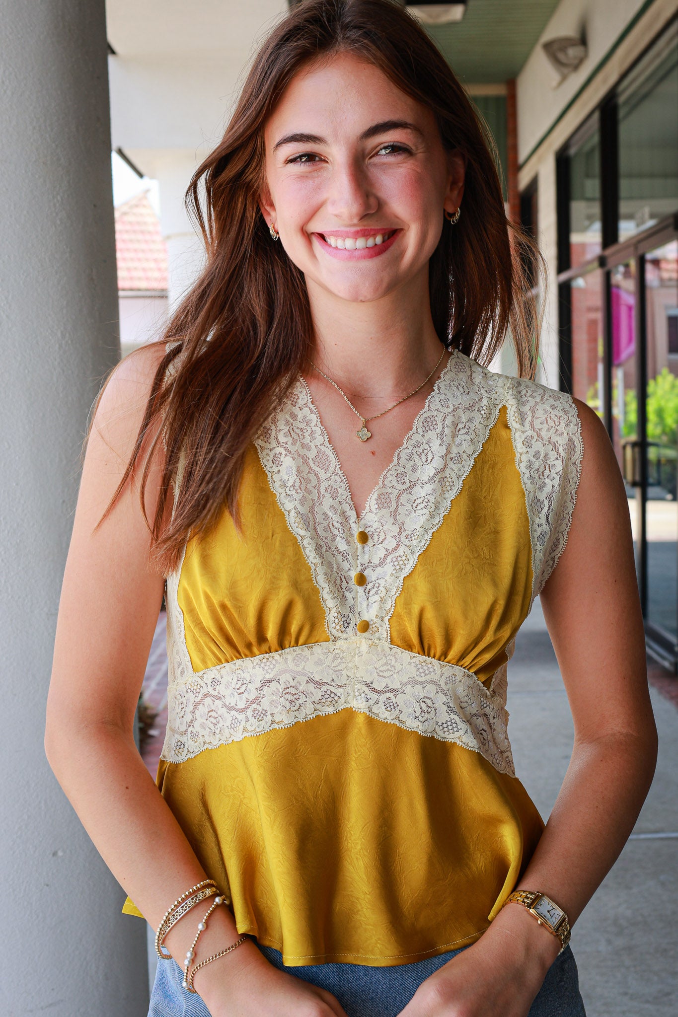 A smiling young woman wearing a sleeveless mustard yellow top with cream lace trim and a buttoned V-neckline, photographed outdoors on a covered walkway.