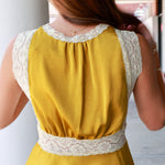 A woman viewed from behind wearing a sleeveless mustard yellow top with cream lace trim at the neckline, underarms, and waist, paired with light blue jeans.