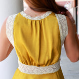 A woman viewed from behind wearing a sleeveless mustard yellow top with cream lace trim at the neckline, underarms, and waist, paired with light blue jeans.