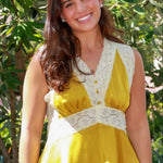 A woman wearing a sleeveless yellow and white lace peplum top with a denim mini skirt, standing outdoors in front of green foliage.