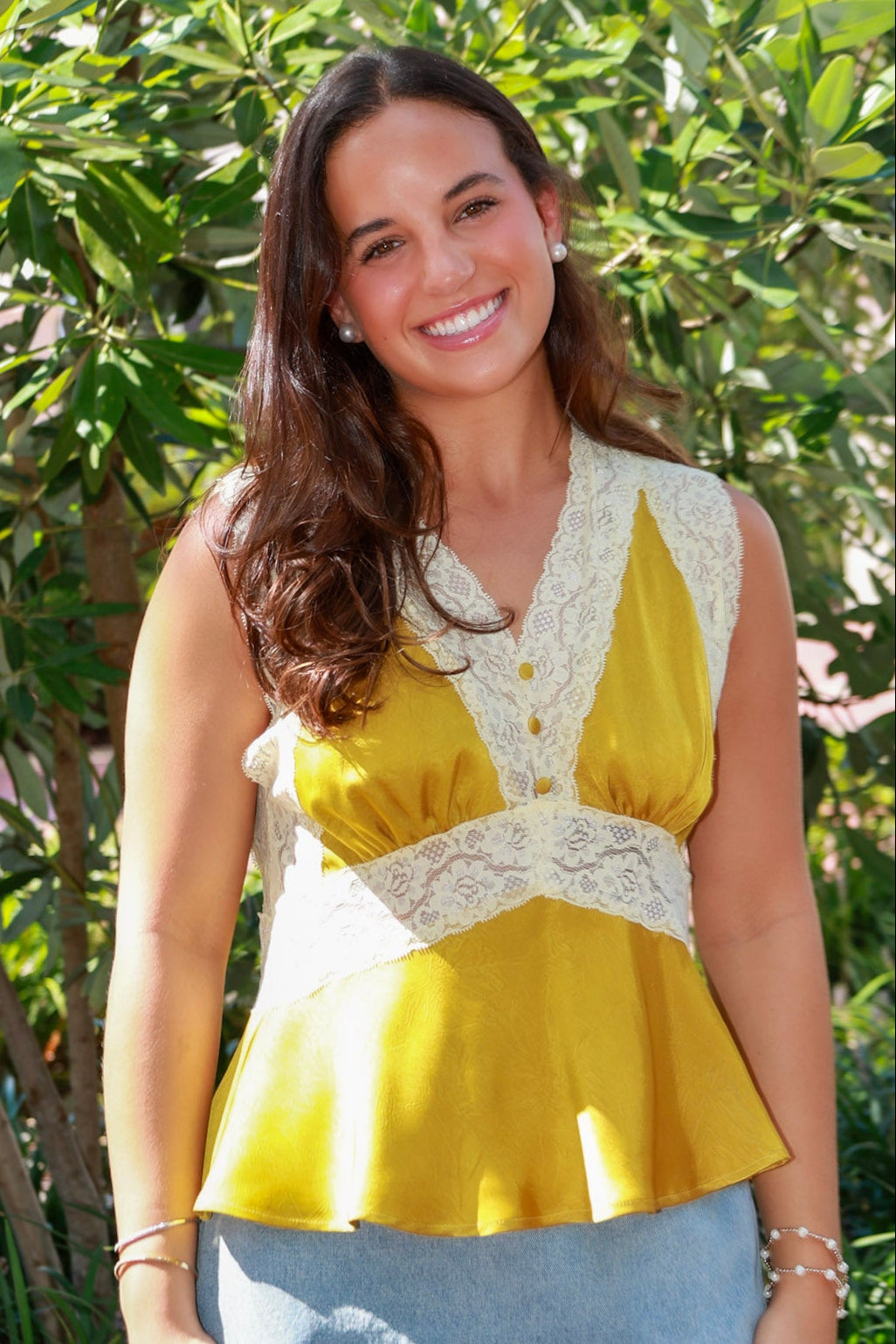 A woman wearing a sleeveless yellow and white lace peplum top with a denim mini skirt, standing outdoors in front of green foliage.