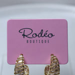 Gold huggie hoop earrings with textured horizontal ridges and small clear stones, displayed on a pink card labeled 'Rodéo BOUTIQUE'.