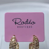 Gold huggie hoop earrings with textured horizontal ridges and small clear stones, displayed on a pink card labeled 'Rodéo BOUTIQUE'.