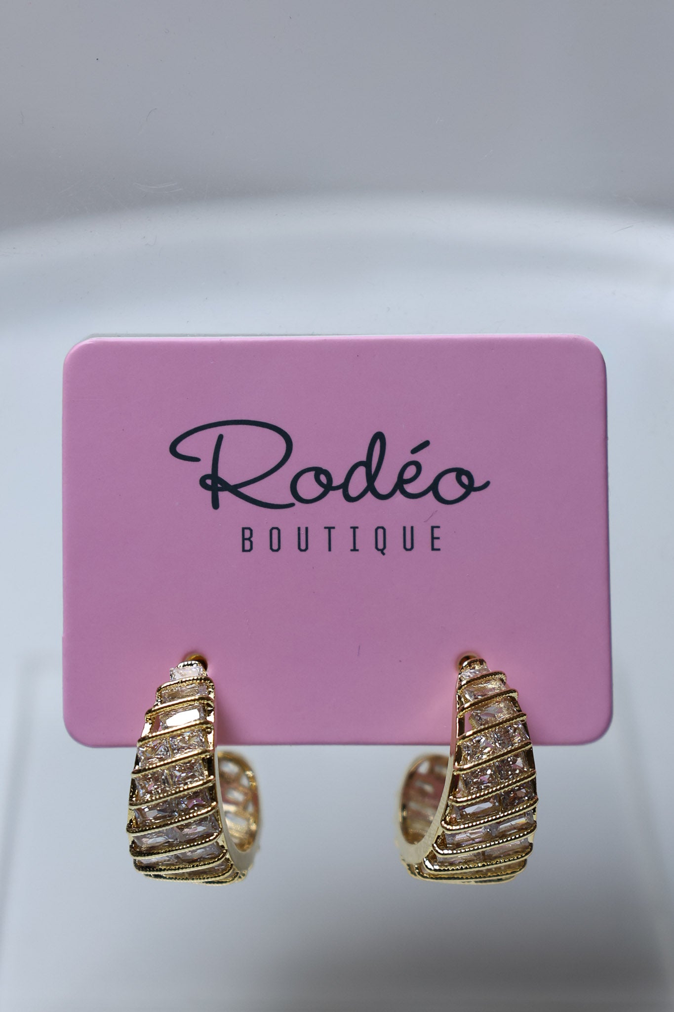 Gold huggie hoop earrings with textured horizontal ridges and small clear stones, displayed on a pink card labeled 'Rodéo BOUTIQUE'.