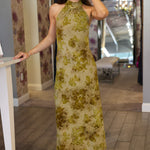 Woman wearing a green floral velvet dress standing in a room with patterned walls and a mirror.