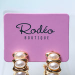 A pair of gold hoop earrings with stacked faux-pearl beads, displayed on a pink card labeled 'Rodéo BOUTIQUE' against a light blue background.
