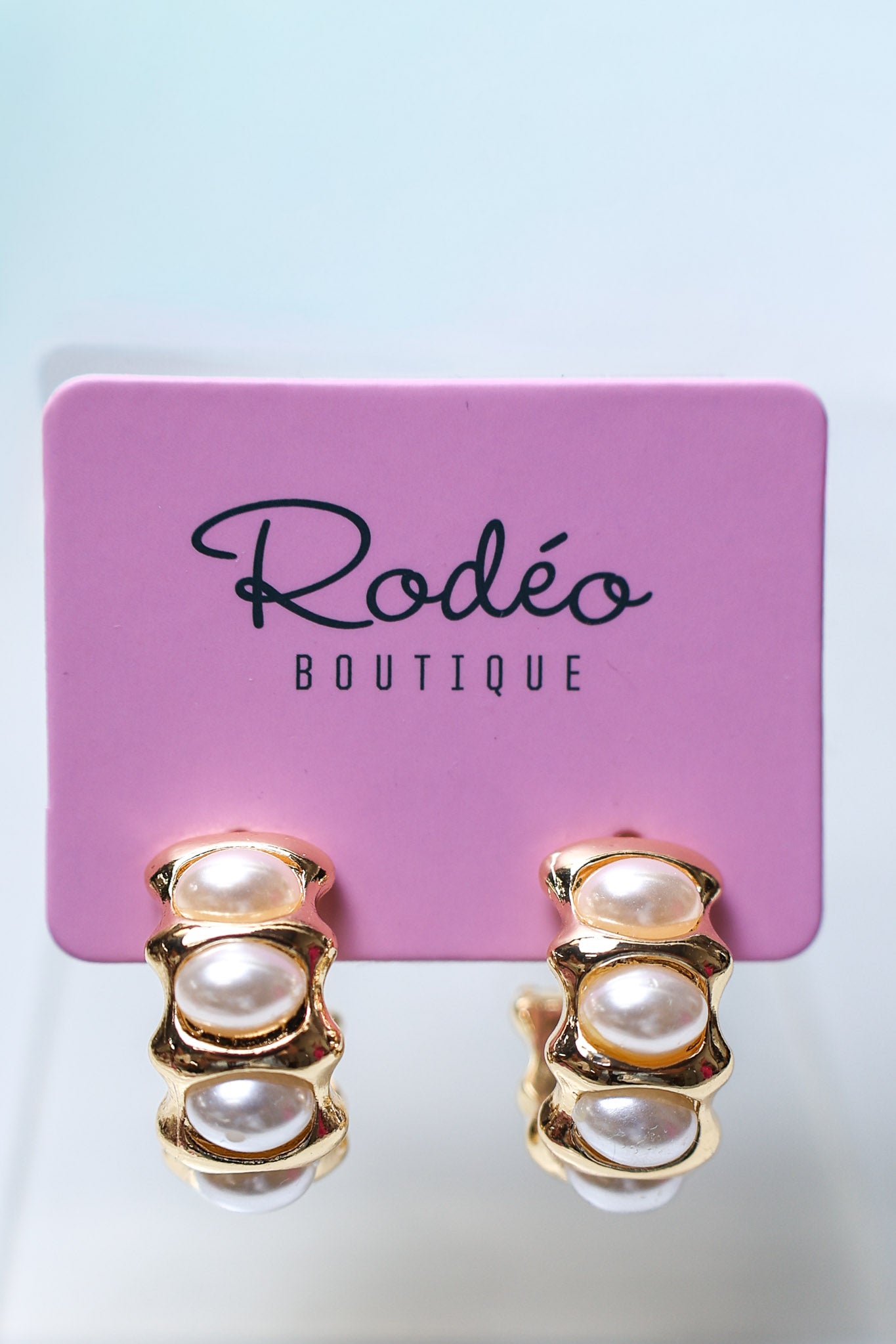 A pair of gold hoop earrings with stacked faux-pearl beads, displayed on a pink card labeled 'Rodéo BOUTIQUE' against a light blue background.