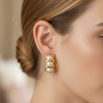 A close-up side profile of a woman wearing a gold-toned triple-row pearl stud earring, with her hair in a low bun against a softly blurred indoor background.